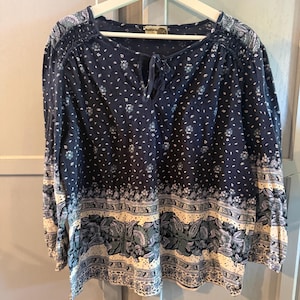 May include: A long-sleeved navy blue blouse featuring a white and light blue floral pattern. The garment has a round neckline with a tie closure, gathered shoulder detailing, and a wide horizontal band of intricate botanical print at the hem.