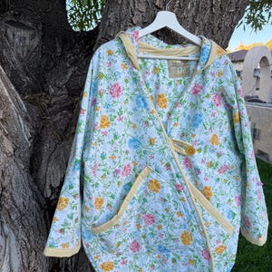 May include: A white hooded jacket featuring a colorful floral pattern with pink, yellow, and blue flowers on green vines. The jacket has a wrap-style front closure with a single yellow button, long sleeves, and yellow trim along the edges.