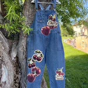 May include: Blue denim overalls featuring multiple cherry-themed patches and graphics. The chest patch displays a bottle with the text Cherry Cola, while the leg patches feature various cherry illustrations and the text Cherry Sweet.