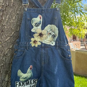 May include: Blue denim overalls featuring a rooster graphic, floral appliques, and a lower left leg print that reads FARMERS MARKET. The garment has a white ruffled hem patterned with various chicken illustrations and is displayed on a white hanger.