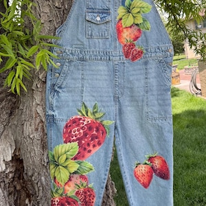 May include: Light blue denim overalls featuring a hand-painted strawberry design. The garment includes a chest pocket, side pockets, and adjustable shoulder straps. Vibrant red strawberries with green leaves are painted on the chest and legs.