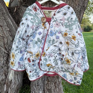 May include: A quilted hooded jacket featuring a white base with a colorful floral print of daisies, lilies, and various wildflowers. The jacket has a dark red trim, a single large wooden button closure at the neck, and a large front pocket.