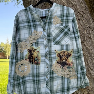 May include: A long-sleeve button-down shirt in a green and white plaid pattern. The shirt features two printed images of Highland cattle surrounded by flowers, accented with beige lace appliques on the front panels and a chest pocket.