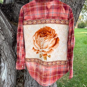May include: A long-sleeve button-down shirt featuring a red and orange plaid pattern with a bleached effect. The back panel displays a large, detailed orange peony flower graphic on a cream-colored background with decorative border trim.