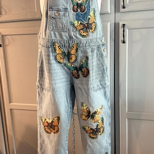May include: Light blue denim overalls featuring a series of colorful butterfly prints scattered across the chest, waist, and legs. The garment includes a front bib pocket, adjustable shoulder straps with metal hardware, and side pockets.