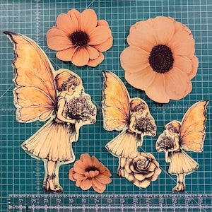 May include: Three die-cut paper fairy figures with large wings holding bouquets and three peach-colored flowers. A ruler at the bottom shows the scale in inches. The fairies wear dresses and have detailed line art, arranged on a green grid mat.