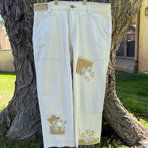 May include: Off-white denim pants featuring a high-waisted cut, a button closure, and visible stitching. The pants are decorated with several rectangular tan fabric patches, each adorned with a small, colorful floral embroidery design.