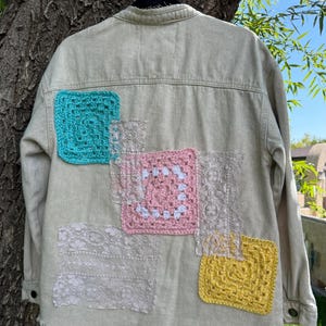 May include: Beige denim jacket with colorful crochet and lace patches. The back of the jacket features turquoise, pink, yellow, and white crochet squares, along with lace patches. The jacket has a frayed hem and button closures.