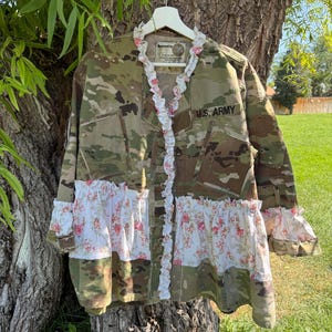 May include: A camouflage jacket featuring a white ruffled trim with a pink floral pattern along the front opening, hem, and cuffs. The jacket includes a black U.S. ARMY patch on the chest and is displayed on a white hanger against a tree.