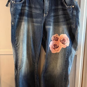 May include: Dark blue denim jeans featuring a faded wash, silver-tone hardware, and a floral print of pink roses on the upper left thigh and lower right leg. The jeans have a classic five-pocket design with visible stitching and a button closure.