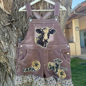 May include: Brown denim overall shorts with a cow graphic on the front pocket. The shorts have a floral print trim at the hem and the word "FARM" printed on the front. A cow and the word "MOO" are also printed on the shorts.