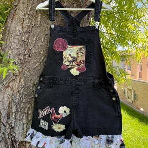 May include: Black denim overalls featuring a chest pocket with a rooster and rose graphic, a leg patch with a rooster and the text Farm Eggs, and a white ruffled hem patterned with small chickens and floral motifs, suspended on a white hanger.