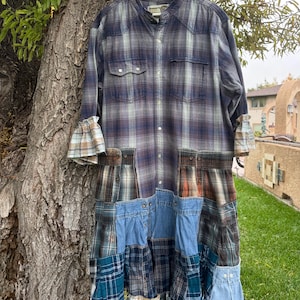 May include: A long-sleeved shirt dress constructed from a patchwork of various plaid flannel and denim fabrics. The garment features a mandarin collar, a button-down front, two chest pockets, and ruffled sleeve cuffs with a multi-layered hem.