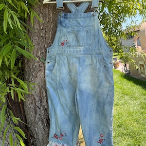 May include: Light blue denim overalls featuring a faded wash, a front bib pocket, and adjustable shoulder straps. The garment is decorated with embroidered red and pink floral motifs on the bib and leg cuffs, which are finished with white ruffled trim.