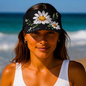 May include: A black visor featuring an embroidered design of two white daisy flowers with yellow centers, surrounded by green leaves and small white buds. The embroidery is centered on the front panel of the visor, which has a curved brim.