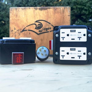 May include: A black box with a red digital display showing 119V, 0.0A, and 60. A black electrical outlet with two white outlets and two USB ports. A white electrical plug with five prongs. A wooden box with the word "Joyce" written on it.