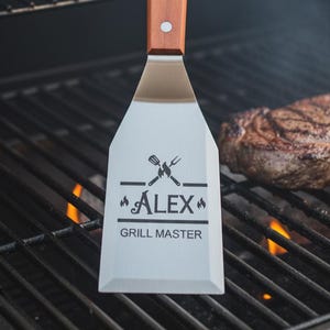 May include: A metal spatula with a wooden handle secured by two rivets. The flat blade features a laser-engraved design with a crossed spatula and fork icon, the name ALEX, and the text GRILL MASTER centered above a horizontal line.