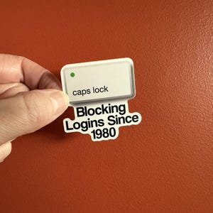 May include: A white sticker with the words "caps lock" and "Blocking Logins Since 1980" in black text. The sticker has a green dot and is held against an orange background.
