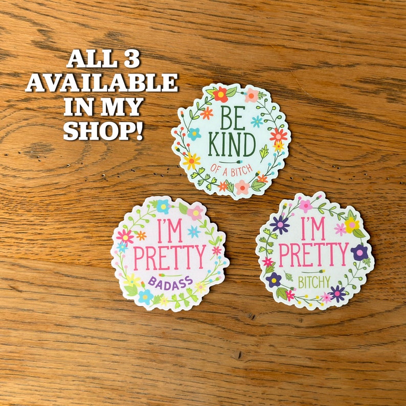 May include: Three round floral stickers with different sayings. The first sticker says "Be Kind of a Bitch" in a white font with a pink outline. The second sticker says "I'm Pretty Badass" in a white font with a pink outline. The third sticker says "I'm Pretty Bitchy" in a white font with a pink outline.