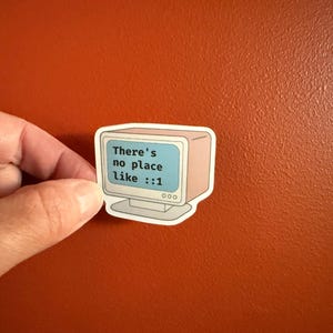May include: A sticker with a vintage computer monitor design. The screen shows the text "There's no place like ::1" in blue. The sticker has a light pink border and a white background, suitable for laptops or notebooks.
