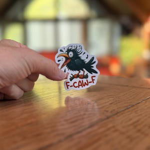 May include: A sticker featuring a cartoon crow with an open beak and the text "F-CAW-F". The crow is black with a yellow beak and is perched on a brown branch. The sticker is white with a clear border.