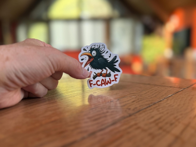 May include: A sticker featuring a cartoon crow with an open beak and the text "F-CAW-F". The crow is black with a yellow beak and is perched on a brown branch. The sticker is white with a clear border.