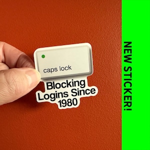 May include: A white sticker with the words "caps lock" and "Blocking Logins Since 1980" in black text. The sticker has a green dot and is held against an orange background. A green vertical stripe on the right says "NEW STICKER!"