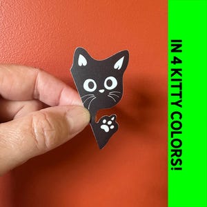 May include: A black cat sticker with white accents, featuring large eyes and a paw print. The sticker is held against an orange background. Text on the right side reads "IN 4 KITTY COLORS!"