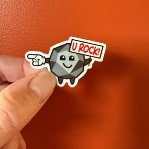 以下が含まれることがあります： 笑顔の顔をした灰色の漫画の岩のキャラクターが、「U ROCK！」と書かれた看板を持っています。岩のキャラクターは左手を左に向けています。