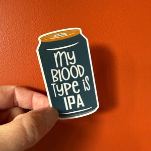 May include: A sticker in the shape of a beer can, navy blue with a gold top. The sticker reads "my BLOOD TYPE is IPA" in white, bold lettering. The sticker is held against an orange background.