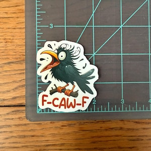 May include: A sticker featuring a cartoon crow with a wide open beak and surprised expression. The crow is perched on a branch, with the text "F-CAW-F" in red. The sticker is on a gray cutting mat with inch measurements.