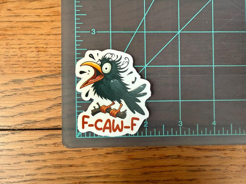May include: A sticker featuring a cartoon crow with a wide open beak and surprised expression. The crow is perched on a branch, with the text "F-CAW-F" in red. The sticker is on a gray cutting mat with inch measurements.