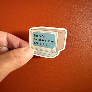 Puede incluir: Una pegatina con un diseño de ordenador retro con el texto "There's no place like 127.0.0.1" en la pantalla. La pegatina se sostiene sobre un fondo naranja.