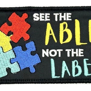 May include: Black embroidered patch with a puzzle piece design in red, yellow, blue, and light blue. The text "See the Able Not the Label" is embroidered in white and yellow.
