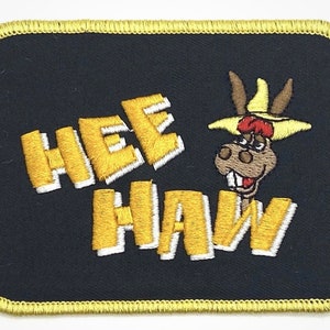 May include: A black embroidered patch with a yellow border. The patch features a cartoon donkey wearing a yellow hat and the words "Hee Haw" in yellow with white outlines.