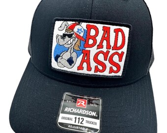 May include: Black trucker hat with a red, white, and blue patch that says "BAD ASS" and features a cartoon dog wearing sunglasses. The hat is adjustable and has a Richardson logo.