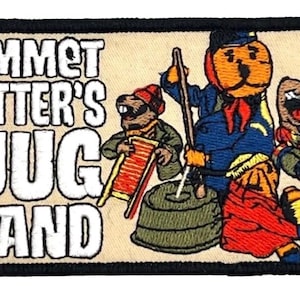 Puede incluir: Parche de Emmet Otter's Jug Band con borde negro. El parche presenta un fondo beige con el nombre de la banda en letras blancas. Nutrias de dibujos animados tocan instrumentos con trajes coloridos.