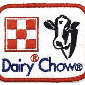 以下が含まれることがあります： 赤と白のチェック柄のロゴに、白黒の牛のイラストと「Dairy Chow」の文字、登録商標のシンボルが描かれています。