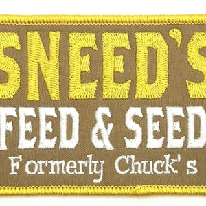 Może przedstawiać: Naszywka haftowana w kolorze brązowym i żółtym z tekstem "Sneed's Feed & Seed" w białym i żółtym kolorze. Tekst "Formerly Chuck's" również jest w kolorze białym.