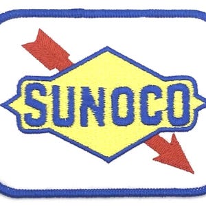 May include: A white square patch with a blue border. Inside the square is a yellow diamond with a blue "SUNOCO" logo. A red arrow points diagonally across the diamond.