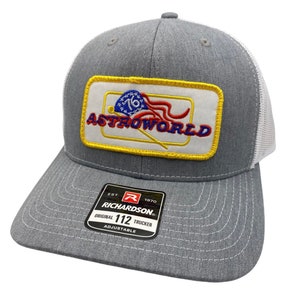 May include: A grey and white trucker hat with a yellow, red, white and blue embroidered patch that says "Astroworld" and features a baseball bat and an American flag. The hat has a black label that says "Richardson" and "Original 112 Trucker Adjustable".