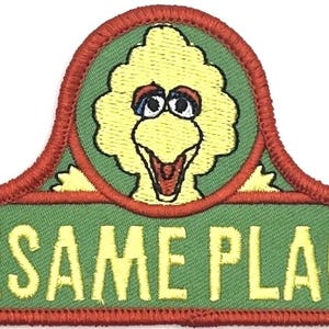 Può includere: Una toppa ricamata che presenta il logo di Sesame Place con il testo SESAME PLACE in lettere gialle su sfondo verde. Sopra il testo si trova un emblema circolare contenente un personaggio giallo Big Bird con dettagli arancioni e neri.