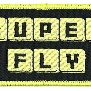 May include: A black fabric patch with yellow stitching. The patch has the words "SUPER FLY" written in yellow block letters.