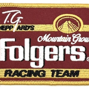 May include: Embroidered patch with a maroon background and gold trim. The patch features the text "T.G. Sheppard's" and "Folgers" in white, with "Mountain Grown" and "Racing Team" in black. A mountain graphic is also present.