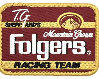 May include: Embroidered patch with a maroon background and gold trim. The patch features the text "T.G. Sheppard's" and "Folgers" in white, with "Mountain Grown" and "Racing Team" in black. A mountain graphic is also present.