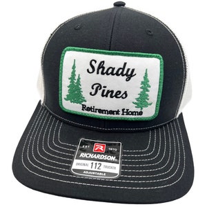 Puede incluir: Gorra de camionero negra y blanca con un parche verde que dice "Shady Pines Retirement Home" con dos pinos verdes. La gorra tiene una parte trasera de malla blanca y una parte delantera negra con una etiqueta Richardson 112.
