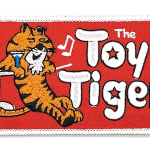 May include: A red embroidered patch with a white border featuring a cartoon tiger holding a drink and the text "The Toy Tiger" with musical notes on either side.
