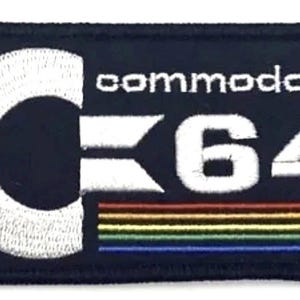 May include: Embroidered patch featuring the Commodore 64 logo. The design includes a white 'C' and the text 'commodore' above '64' in white. A rainbow-colored stripe is at the bottom, all on a navy blue background.