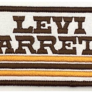 Può includere: Toppa ricamata con la scritta "LEVI GARRETT" in lettere marroni. La toppa ha uno sfondo bianco con strisce marroni e gialle nella parte inferiore e un bordo marrone.
