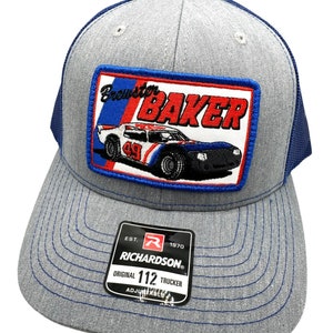 May include: Gray and blue trucker hat with a red, white, and blue patch featuring a race car with the text "Brewster Baker" and the number "49". The hat has a Richardson logo and the text "Original 112 Trucker Adjustable".
